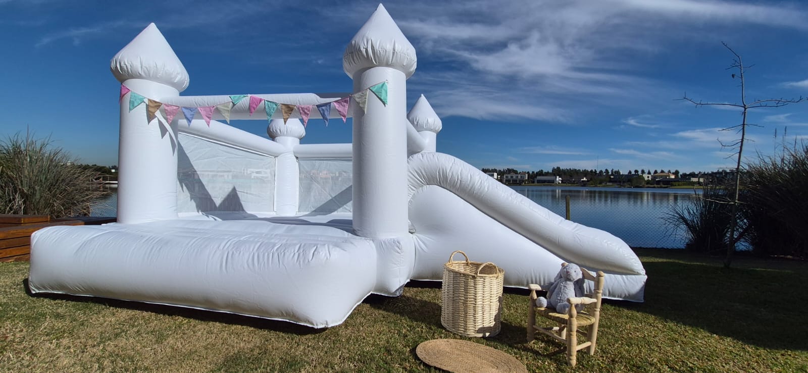 Castillo inflable blanco con torres, banderines pastel y tobogán lateral, en un jardín junto al agua.